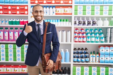 Sticker - Hispanic man with beard working as salesman at pharmacy drugstore smiling happy and positive, thumb up doing excellent and approval sign