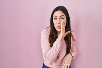 Wall Mural - Young brunette woman standing over pink background hand on mouth telling secret rumor, whispering malicious talk conversation