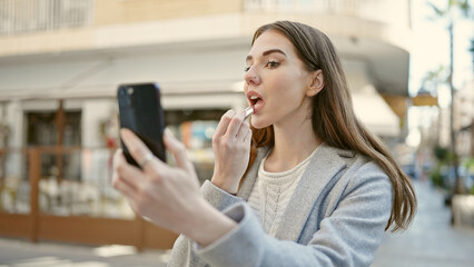 Poster - Young hispanic woman using smartphone as a mirror applying lipstick at coffee shop terrace