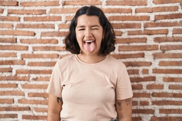 Poster - Young hispanic woman standing over bricks wall sticking tongue out happy with funny expression. emotion concept.