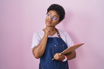 Poster - Young african american woman wearing professional waitress apron holding clipboard with hand on chin thinking about question, pensive expression. smiling with thoughtful face. doubt concept.