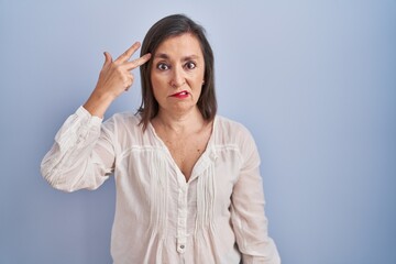 Poster - Middle age hispanic woman standing over blue background shooting and killing oneself pointing hand and fingers to head like gun, suicide gesture.