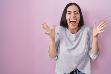 Poster - Young brunette woman standing over pink background celebrating crazy and amazed for success with arms raised and open eyes screaming excited. winner concept