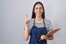 Young Brunette Woman Wearing Professional Waitress Apron And Clipboard Surprised With An Idea Or Question Pointing Finger With Happy Face, Number One