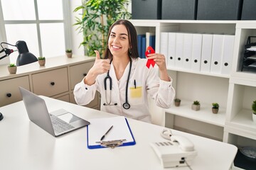 Poster - Hispanic doctor woman holding support red ribbon smiling happy and positive, thumb up doing excellent and approval sign