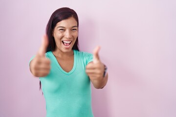 Poster - Young hispanic woman standing over pink background approving doing positive gesture with hand, thumbs up smiling and happy for success. winner gesture.