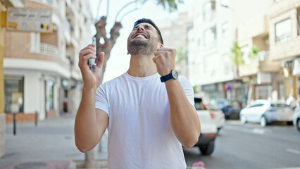 Poster - Young hispanic man using smartphone with winner expression at street