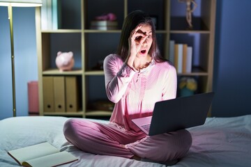 Canvas Print - Young hispanic woman using computer laptop on the bed doing ok gesture shocked with surprised face, eye looking through fingers. unbelieving expression.