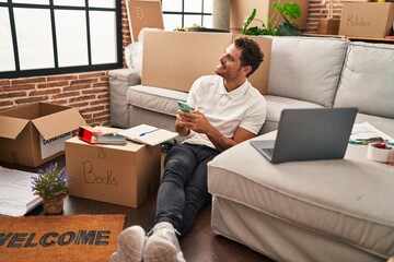 Wall Mural - Young hispanic man using smartphone sitting on floor at new home