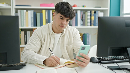 Wall Mural - Young hispanic man student using smartphone writing notes at library university