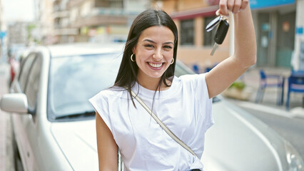 Young beautiful hispanic woman holding key standing by car at street