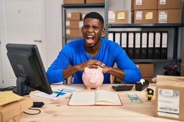 Wall Mural - African american man working at small business ecommerce with piggy bank angry and mad screaming frustrated and furious, shouting with anger. rage and aggressive concept.