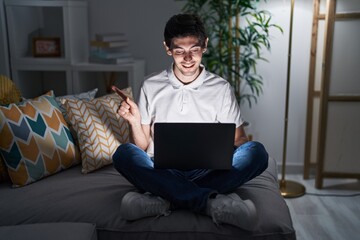 Canvas Print - Young hispanic man using laptop at home at night cheerful with a smile on face pointing with hand and finger up to the side with happy and natural expression