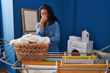 Sticker - Young asian woman hanging clothes at clothesline smelling something stinky and disgusting, intolerable smell, holding breath with fingers on nose. bad smell