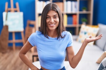 Poster - Brunette young woman sitting on the sofa at home smiling cheerful presenting and pointing with palm of hand looking at the camera.