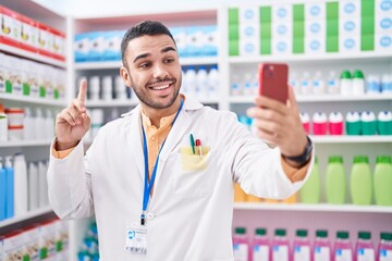 Wall Mural - Young hispanic man working at pharmacy drugstore doing video call with smartphone smiling with an idea or question pointing finger with happy face, number one