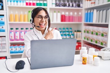 Wall Mural - Young arab woman working at pharmacy drugstore using laptop with a big smile on face, pointing with hand and finger to the side looking at the camera.