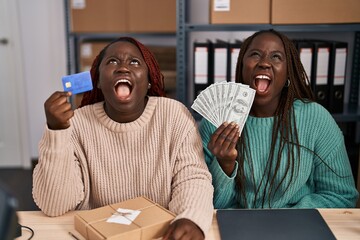 Poster - Two african women working at small business ecommerce holding credit card and banknotes angry and mad screaming frustrated and furious, shouting with anger looking up.