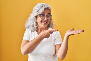 Sticker - Middle age woman with grey hair standing over yellow background amazed and smiling to the camera while presenting with hand and pointing with finger.