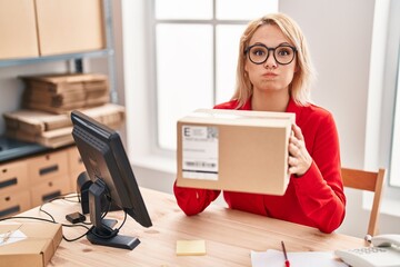 Canvas Print - Blonde woman working at small business ecommerce holding box puffing cheeks with funny face. mouth inflated with air, catching air.