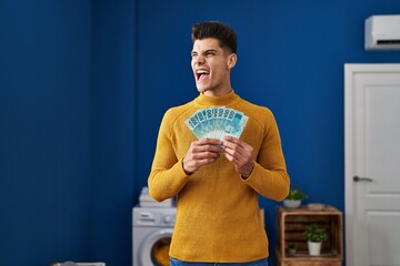 Sticker - Young hispanic man at laundry room holding brazilian reals angry and mad screaming frustrated and furious, shouting with anger. rage and aggressive concept.