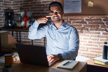 Wall Mural - Hispanic man with beard working at the office at night pointing with hand finger to face and nose, smiling cheerful. beauty concept