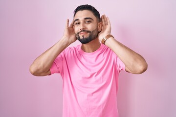 Poster - Hispanic young man standing over pink background trying to hear both hands on ear gesture, curious for gossip. hearing problem, deaf