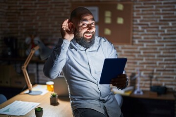 Wall Mural - Young hispanic man with beard and tattoos working at the office at night smiling with hand over ear listening an hearing to rumor or gossip. deafness concept.