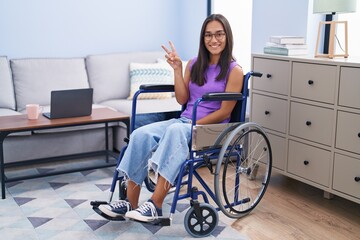 Wall Mural - Young hispanic woman sitting on wheelchair at home showing and pointing up with fingers number two while smiling confident and happy.