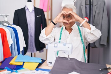 Canvas Print - Middle age man with grey hair dressmaker using sewing machine doing heart shape with hand and fingers smiling looking through sign