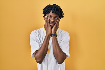 Poster - Young african man with dreadlocks standing over yellow background tired hands covering face, depression and sadness, upset and irritated for problem