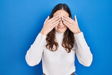 Sticker - Young hispanic woman standing over blue background covering eyes with hands smiling cheerful and funny. blind concept.