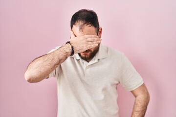 Wall Mural - Plus size hispanic man with beard standing over pink background covering eyes with hand, looking serious and sad. sightless, hiding and rejection concept