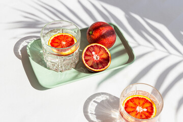 Wall Mural - Creative composition made of half bloody red orange on a tray and two glasses with cocktail on white background with palm tree leaf shadow. Summer refreshment and party concept. Minimal style.