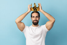 Portrait Of Serious Independent Man With Beard In White T-shirt Wearing Golden Crown, Looking With Arrogance And Confidence, Privileged Status. Indoor Studio Shot Isolated On Blue Background.