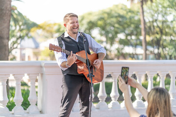 Street musician performing at a park and a girl from the audience taking pictures and filming him