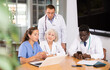 © JackF - In morning meeting, doctors of emergency department confer about patients clinical case. Male health worker with colleagues sitting at table, reading new article on laptop and discussing information