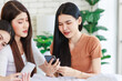 © Bangkok Click Studio - Three Asian pretty cheerful happy female girlfriends in casual outfits sitting on bed smiling chit chat talking discuss holding watching streaming online shows via touchscreen smartphone in bedroom