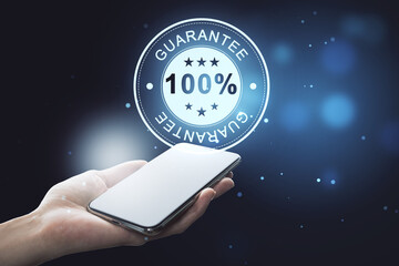 Poster - Close up of female hand holding smartphone with glowing 100% guarantee stamp hologram on dark background with bokeh circles. Quality seal and warranty concept.