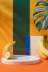Wall Mural - Front view of round podium with ripe banana, green monstera leaf and acrylic sheet arranged on a yellow background. Blank space for display cosmetic or product with ingredient from banana
