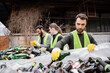 © LIGHTFIELD STUDIOS - Bearded worker in protective gloves and vest putting glass bottle in sack near blurred interracial colleagues in outdoor waste disposal station, garbage sorting and recycling concept