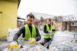 © LIGHTFIELD STUDIOS - Bearded sorter in gloves and protective vest taking glass trash from sack near blurred and smiling multiethnic colleagues in outdoors waste disposal station, garbage sorting and recycling concept