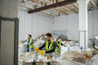© LIGHTFIELD STUDIOS - Male worker in protective vest and gloves holding plastic trash while standing near sacks and blurred interracial colleagues in waste disposal station, garbage sorting and recycling concept
