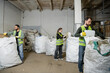 © LIGHTFIELD STUDIOS - Multiethnic workers in high visibility vests and gloves separating trash in sacks while working in waste disposal station, garbage sorting and recycling concept