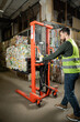 © LIGHTFIELD STUDIOS - Side view of bearded sorter in protective vest and gloves using hand pallet truck with waste paper while working in blurred garbage sorting center, recycling concept