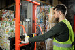 © LIGHTFIELD STUDIOS - Side view of bearded worker in protective vest and glove using hand pallet truck with waste paper while working in blurred garbage sorting center, recycling concept