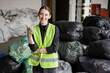 © LIGHTFIELD STUDIOS - Smiling young worker in protective vest and gloves showing like gesture and looking at camera while standing near blurred plastic bags with trash in garbage sorting center, recycling concept