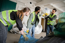 Young Female Worker In Protective Vest And Gloves Holding Plastic Bag Near Blurred Multiethnic Colleagues In Waste Disposal Station, Garbage Sorting And Recycling Concept