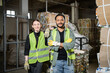 © LIGHTFIELD STUDIOS - Smiling young worker in protective vest and gloves looking at camera near indian colleague crossing arms and standing near waste paper in waste disposal station, garbage sorting and recycling concept