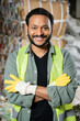 © LIGHTFIELD STUDIOS - Cheerful indian worker in reflective vest and gloves looking at camera and crossing arms while standing near waste paper on blurred background in waste disposal station, garbage recycling concept